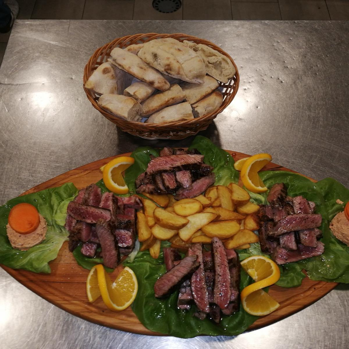 Ramstek narezan na ovalnoj dasci, sa pečenim krompirom, narandžom i korpom lepinja